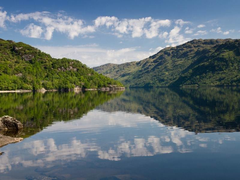 A lake with reflections of green hills in Loch Lomond one of the most popular places to visit in Scotland