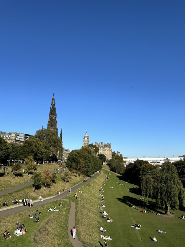 People relaxing on the grass in Princes Street Gardens, Edinburgh, with the Scott Monument and the Balmoral clock tower behind