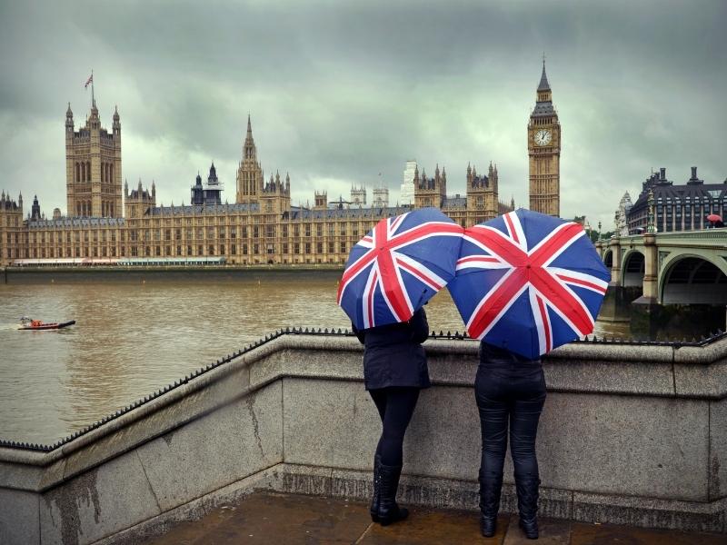 View of 2 people standing with umbrellas opposite the Houses of Parliament in London.