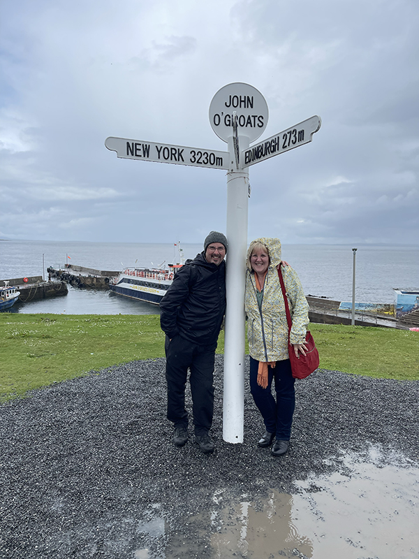 Episode 69 UK Travel planning podcast Doug and Tracy Collins standing beside a sign at John O'Groats.