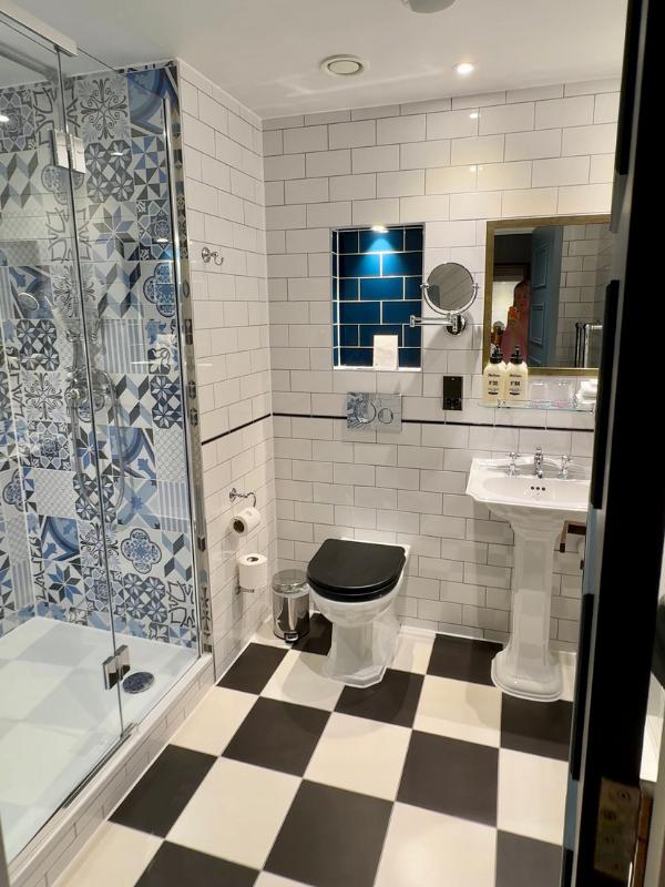 Elegante cuarto de baño de hotel con baldosas monocromáticas en forma de tablero de ajedrez, baldosas blancas del metro, mampara de ducha con motivos azules, lavabo con pedestal y espejo con marco dorado