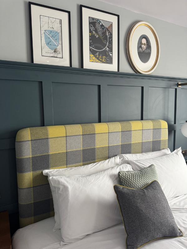 Detalle de la habitación del hotel que muestra una cabecera tapizada en tartán amarillo y gris contra paneles de color verde azulado, con obras de arte enmarcadas con temas de Shakespeare arriba