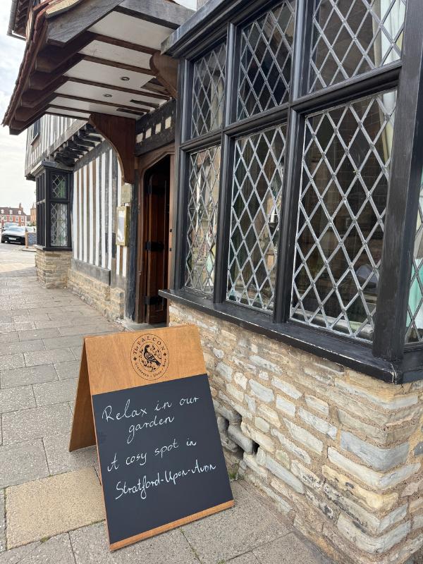 Exterior del Hotel Indigo Stratford-upon-Avon, un edificio histórico con entramado de madera en blanco y negro, ventanas emplomadas y un cartel que dice A-board "Relájese en nuestro jardín, un lugar acogedor en Stratford-upon-Avon"