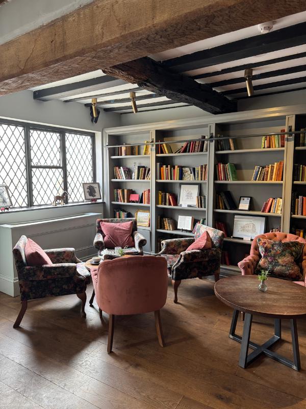 Acogedora sala de lectura con vigas de roble a la vista, ventanas emplomadas, estanterías de piso a techo y sillones florales y de terciopelo que no combinan alrededor de una mesa redonda de madera.