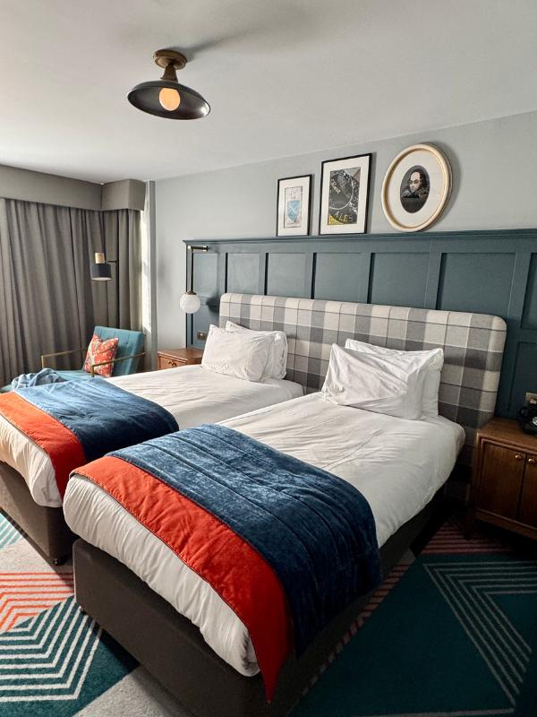 Amplia habitación de hotel con dos camas individuales, cabeceras de tartán gris, paneles de pared verde azulado, corredores de cama azul marino y naranja, una alfombra con motivos geométricos y arte en la pared con temática de Shakespeare.