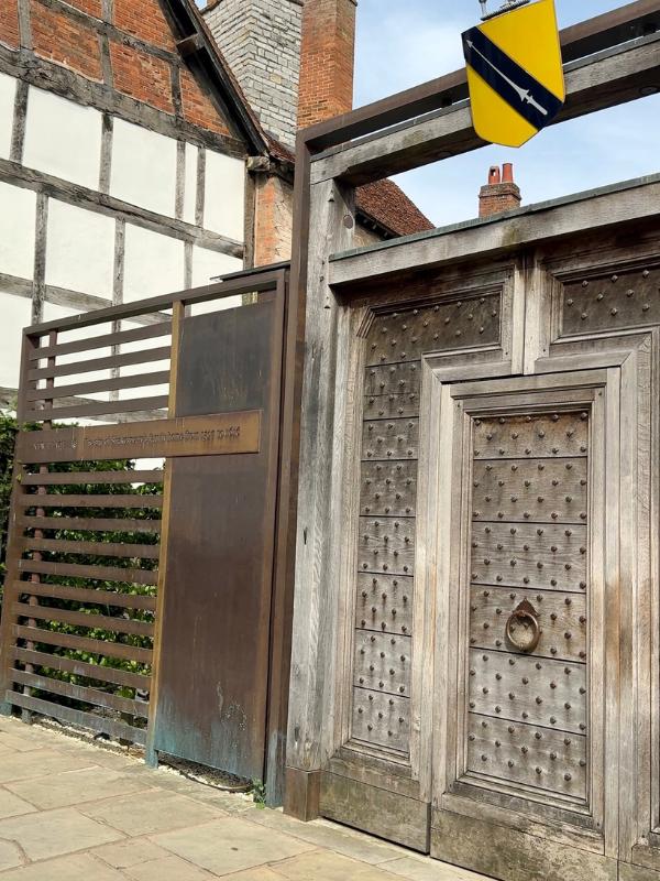 Puerta histórica de madera con clavos y letrero heráldico ornamental que marca la entrada al New Place de Shakespeare, Stratford-upon-Avon, frente a un edificio con entramado de madera en blanco y negro