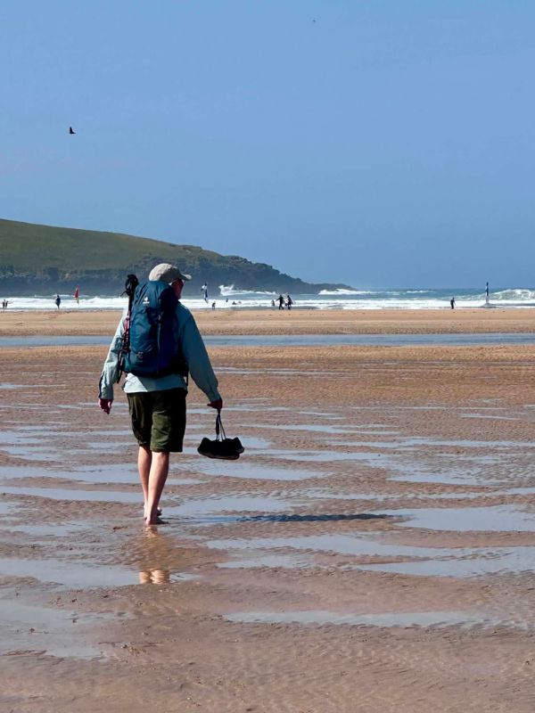 A walker with a backpack crossing wet sand at low tide on a Cornish beach