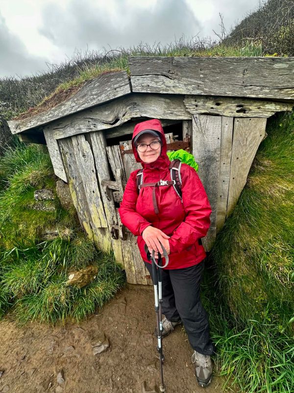 Katherine in a red raincoat with walking poles beside a rustic timber hut on the coast path