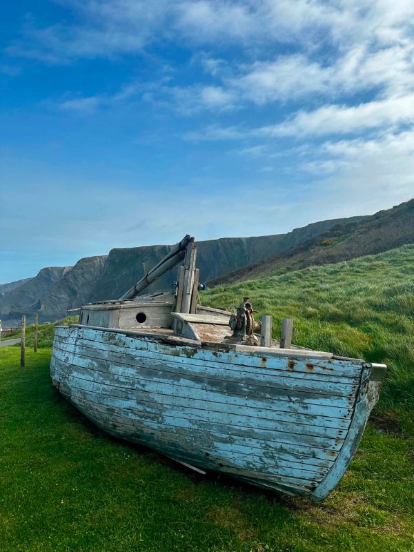 A weathered pale blue wooden fishing boat resting on grass with coastal cliffs behind