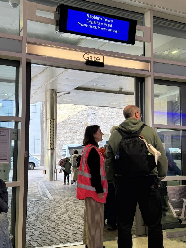 The Rabbie's Tours departure point sign above a gate at Edinburgh Bus Station, with a staff member in a hi-vis vest checking passengers in
