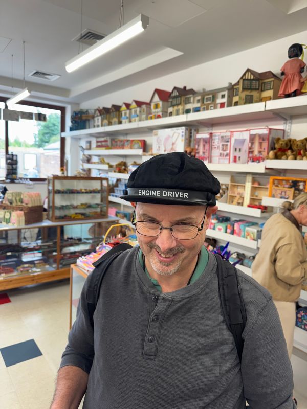 Doug smiling in an Engine Driver cap in the toy shop at Beamish Museum, with dolls' houses on the shelves behind